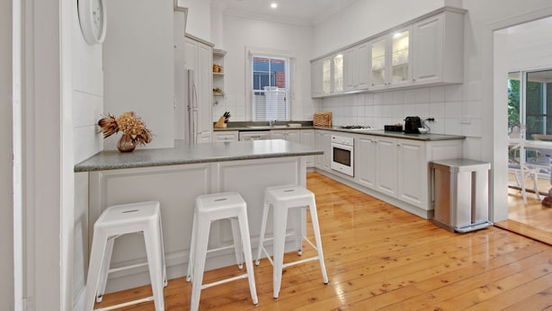 Une cuisine moderne avec des meubles blancs, un îlot central doté de tabourets, une fenêtre avec des stores et une vue sur la verdure à l’extérieur.