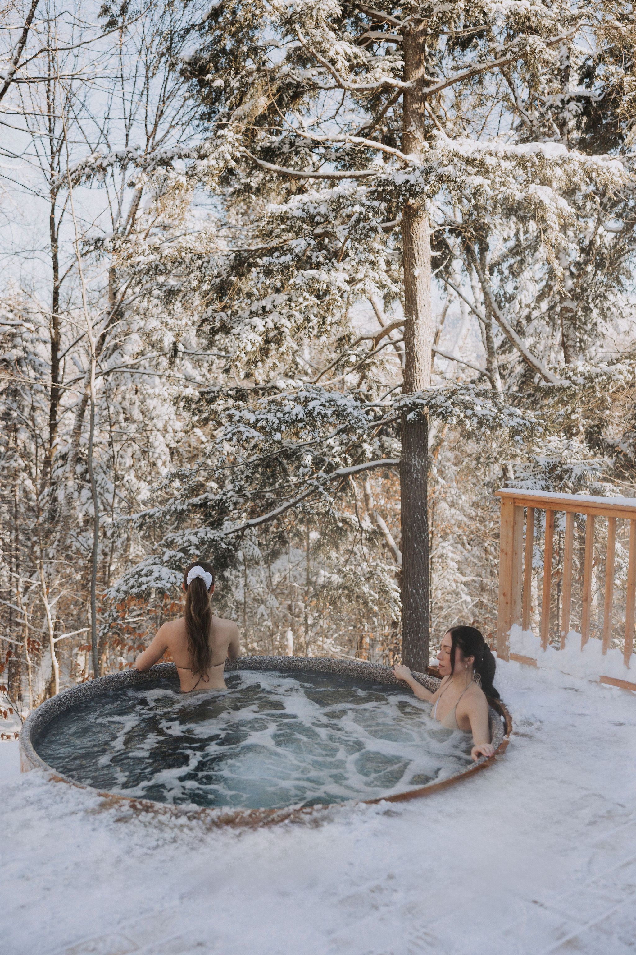 Two individuals in a hot tub surrounded by snow-covered trees.