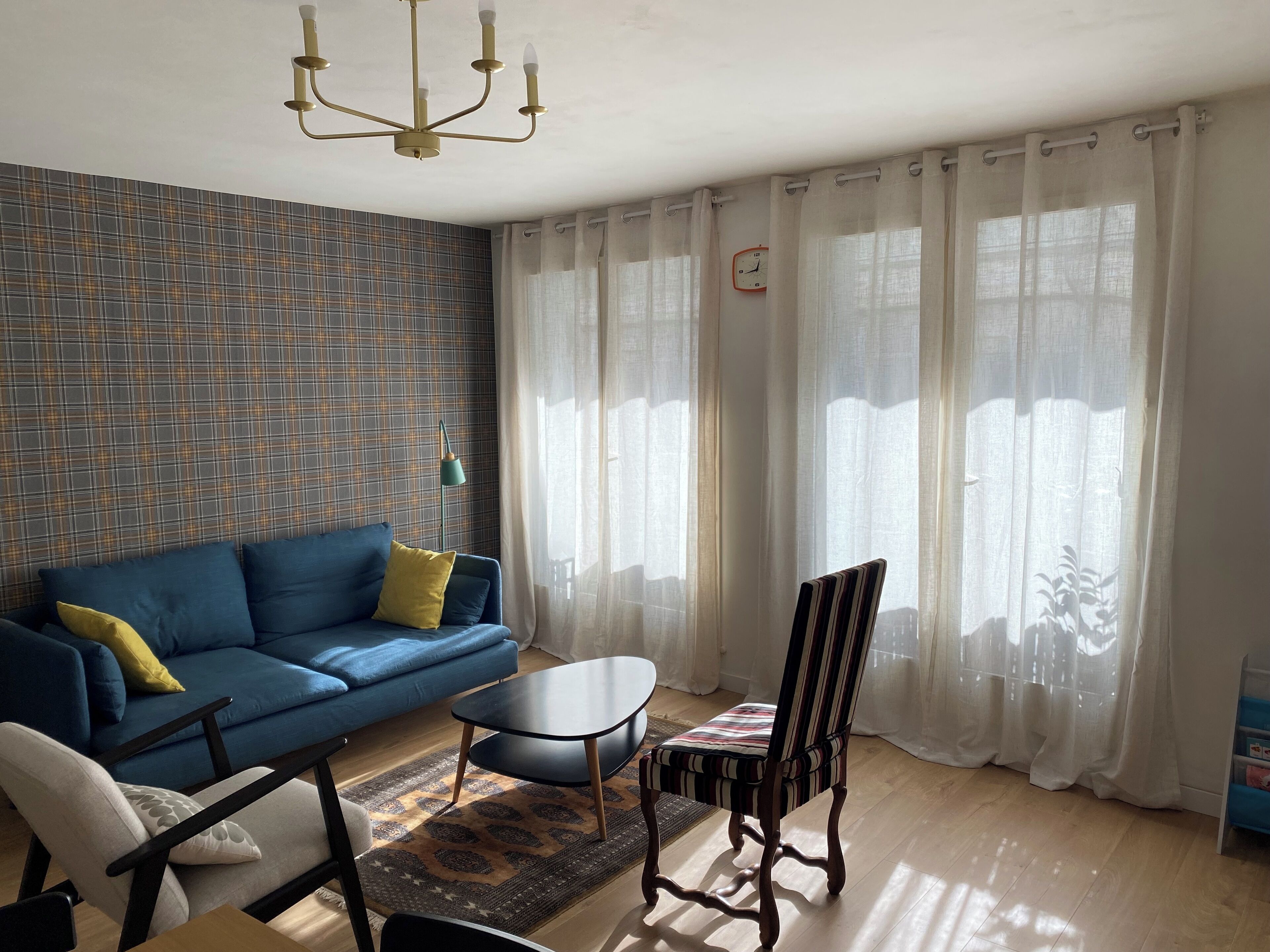 Ein Wohnzimmer mit einem blauen Sofa, einem gelben Kissen, einem gemusterten Teppich, einem Holzstuhl und einem Tisch.