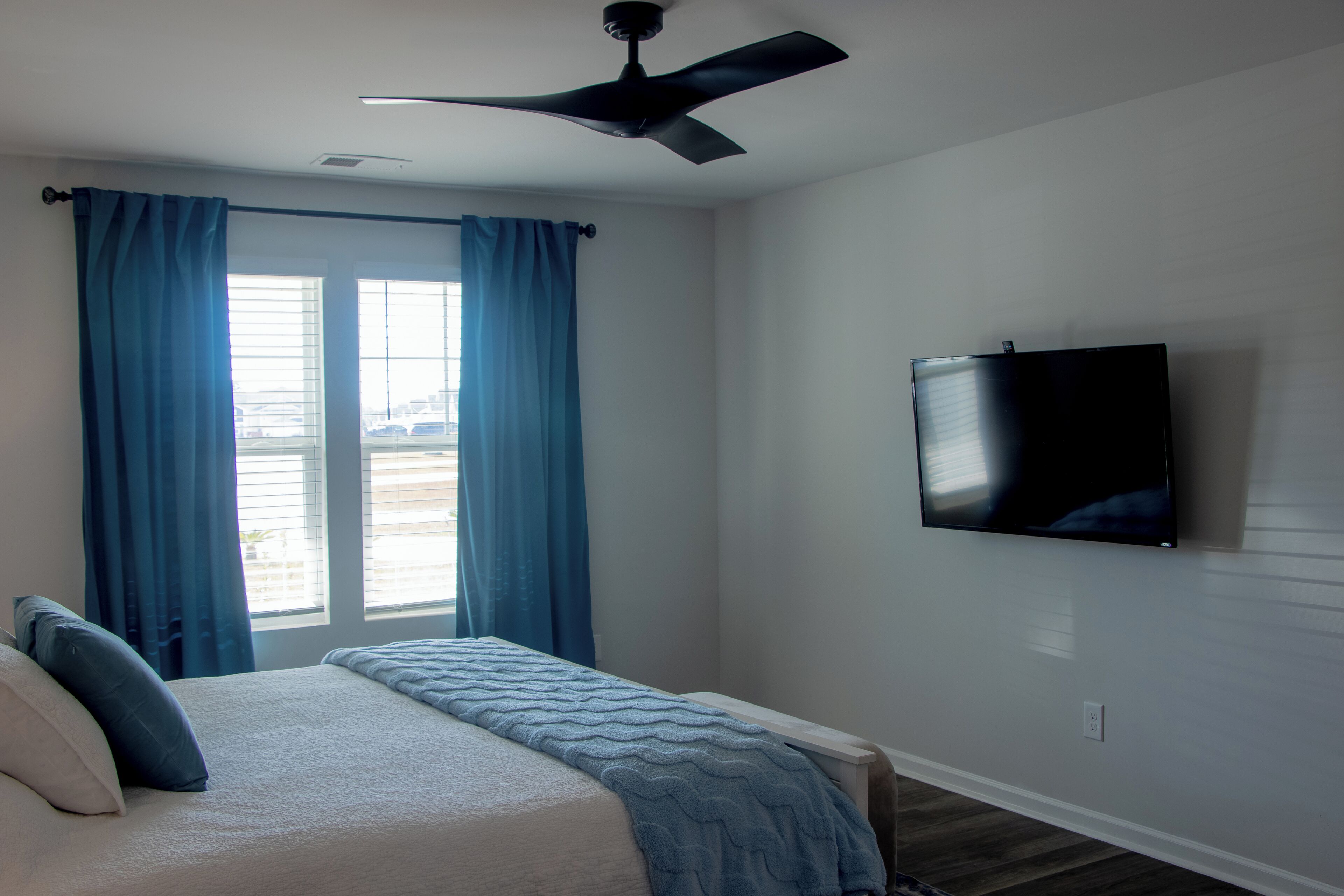 Une chambre à coucher avec un lit, des rideaux, un ventilateur de plafond et un téléviseur fixé au mur.