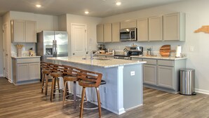 A modern kitchen with grey cabinets, stainless steel appliances, and a central island with bar stools. - New & Modern Home in Southport • Family & Pet Friendly (Southport)