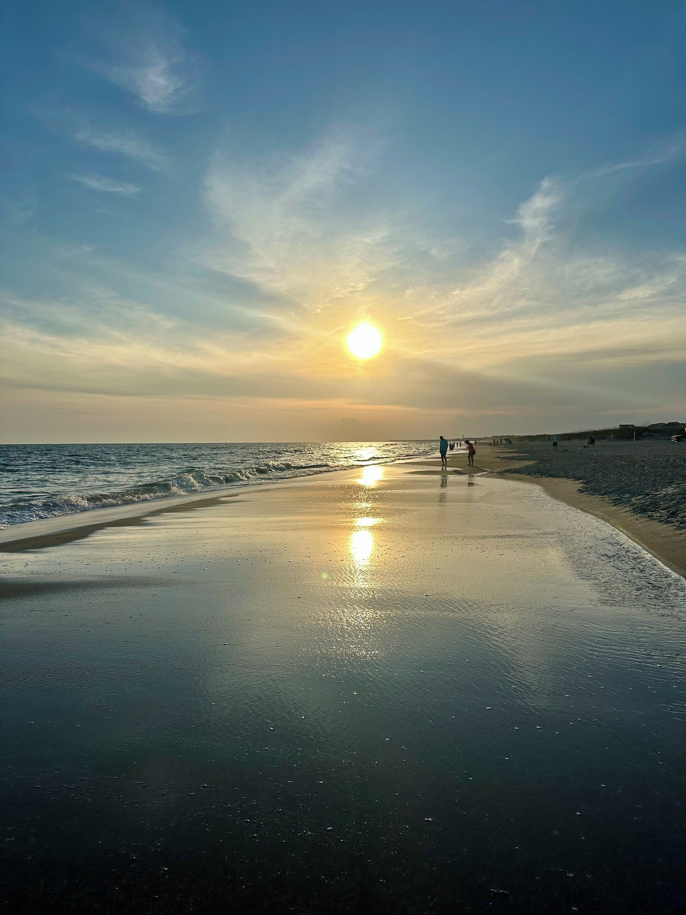 Una playa con un atardecer, mar tranquilo y algunas personas caminando.