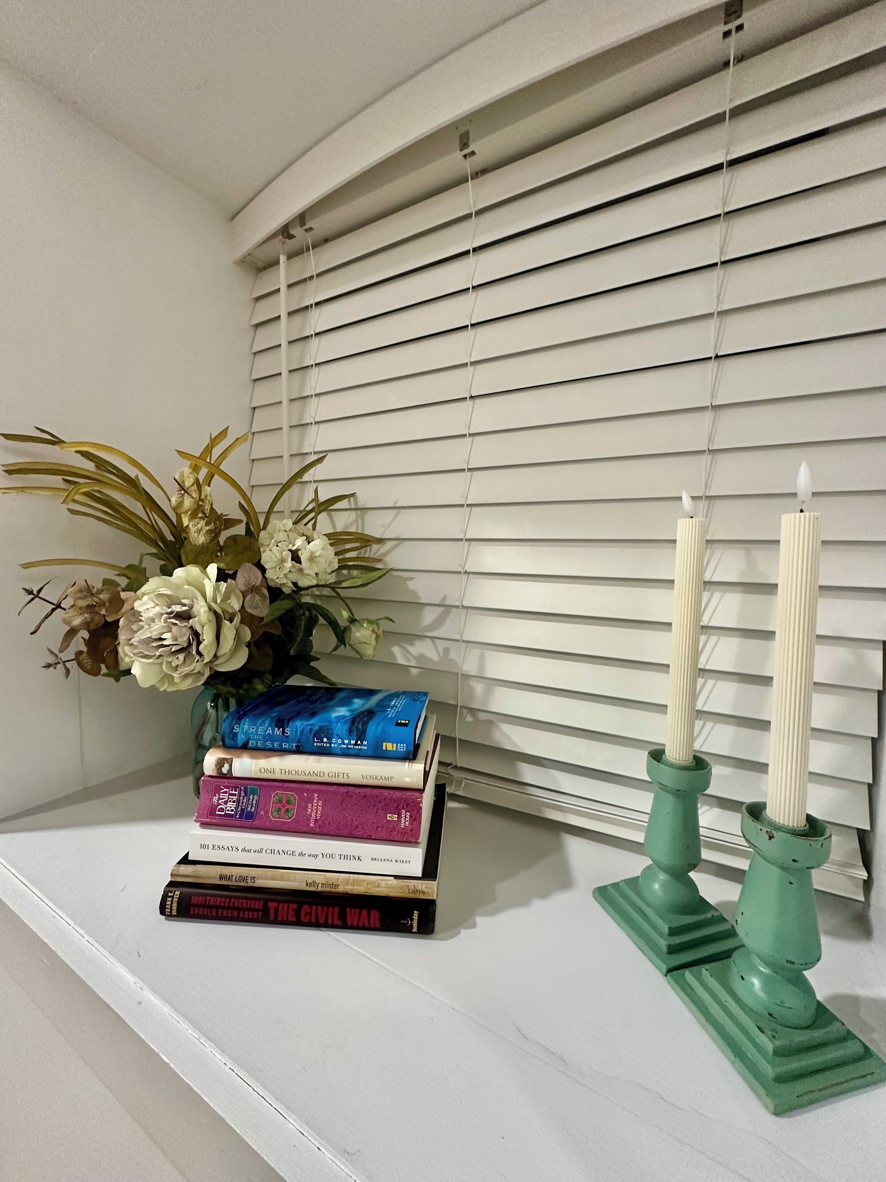 Uno scaffale con libri, un vaso con fiori secchi e due candelabri verdi.