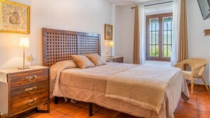 Une chambre à coucher avec une tête de lit en bois, un lit recouvert d’une literie à motifs, une table de chevet avec une lampe, une chaise en osier et une fenêtre avec des rideaux.