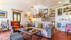 Un salon chaleureux avec un canapé, des fauteuils, une table basse et un coin repas comprenant une table et des chaises. Un escalier mène à l’étage supérieur, et divers objets décoratifs sont exposés sur des étagères et les murs.