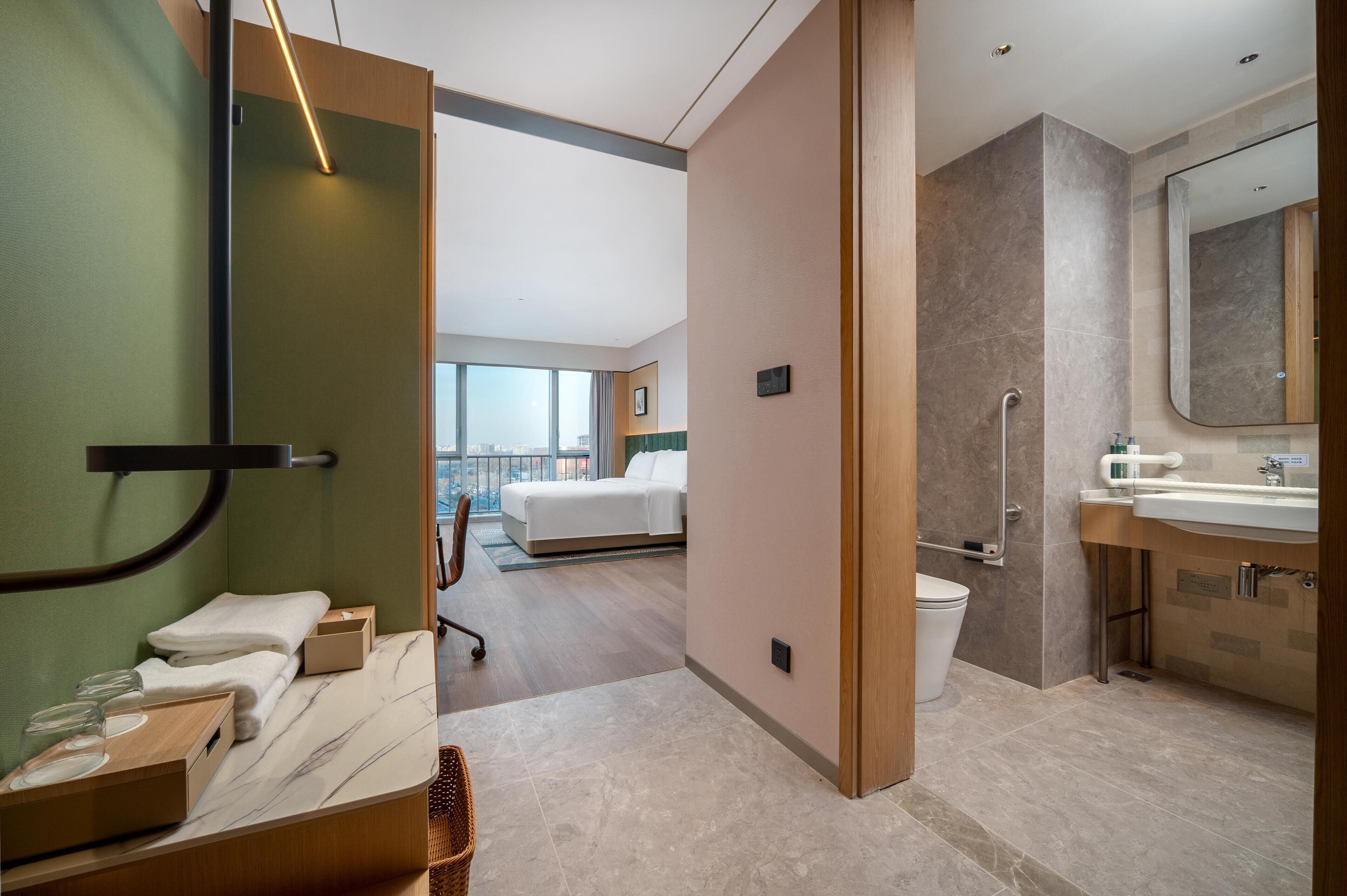 A modern hotel room with a bed, a bathroom with a sink and mirror, and a view of the city through large windows.