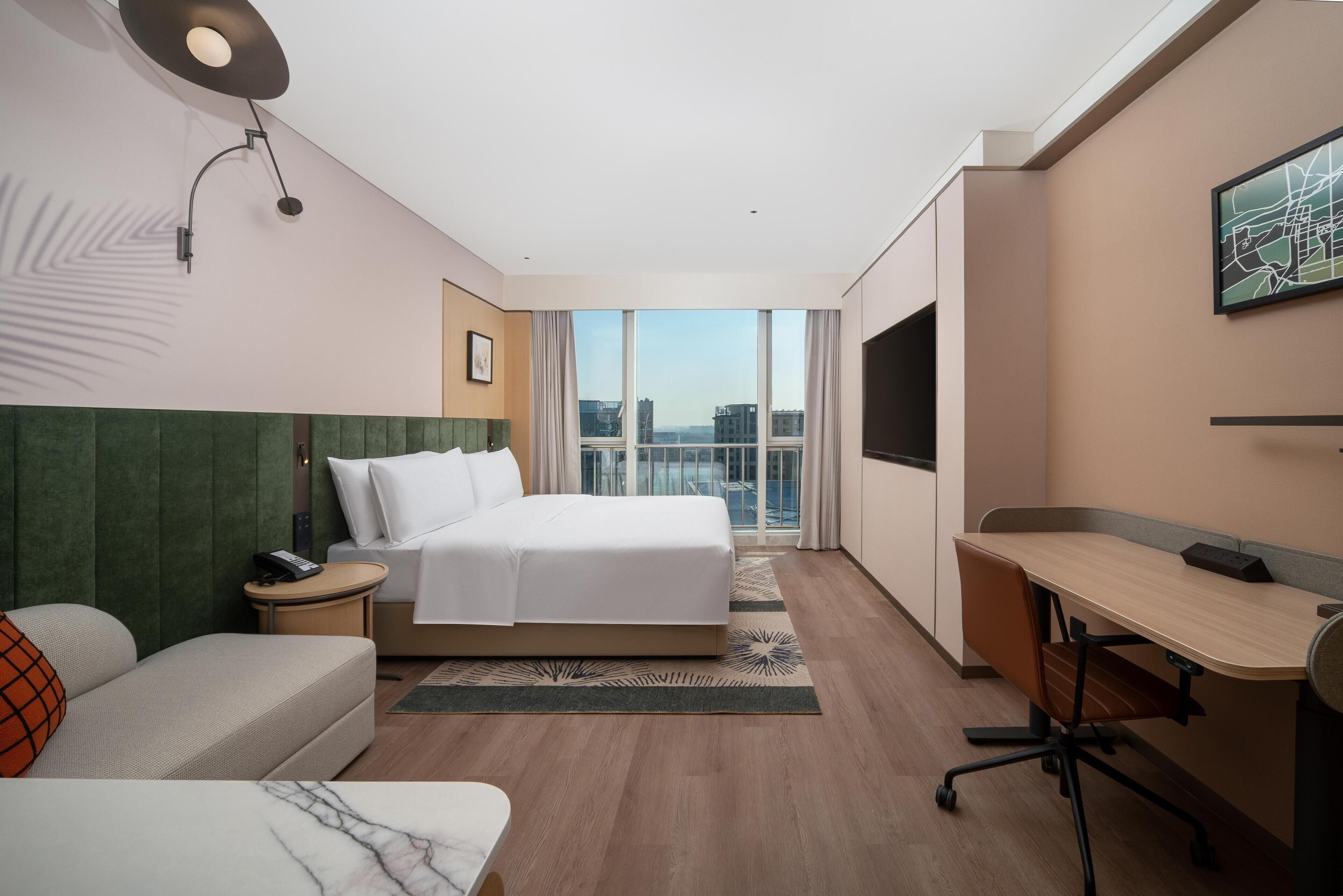 A hotel room with a bed, a desk, a sofa, and a large window offering a city view.
