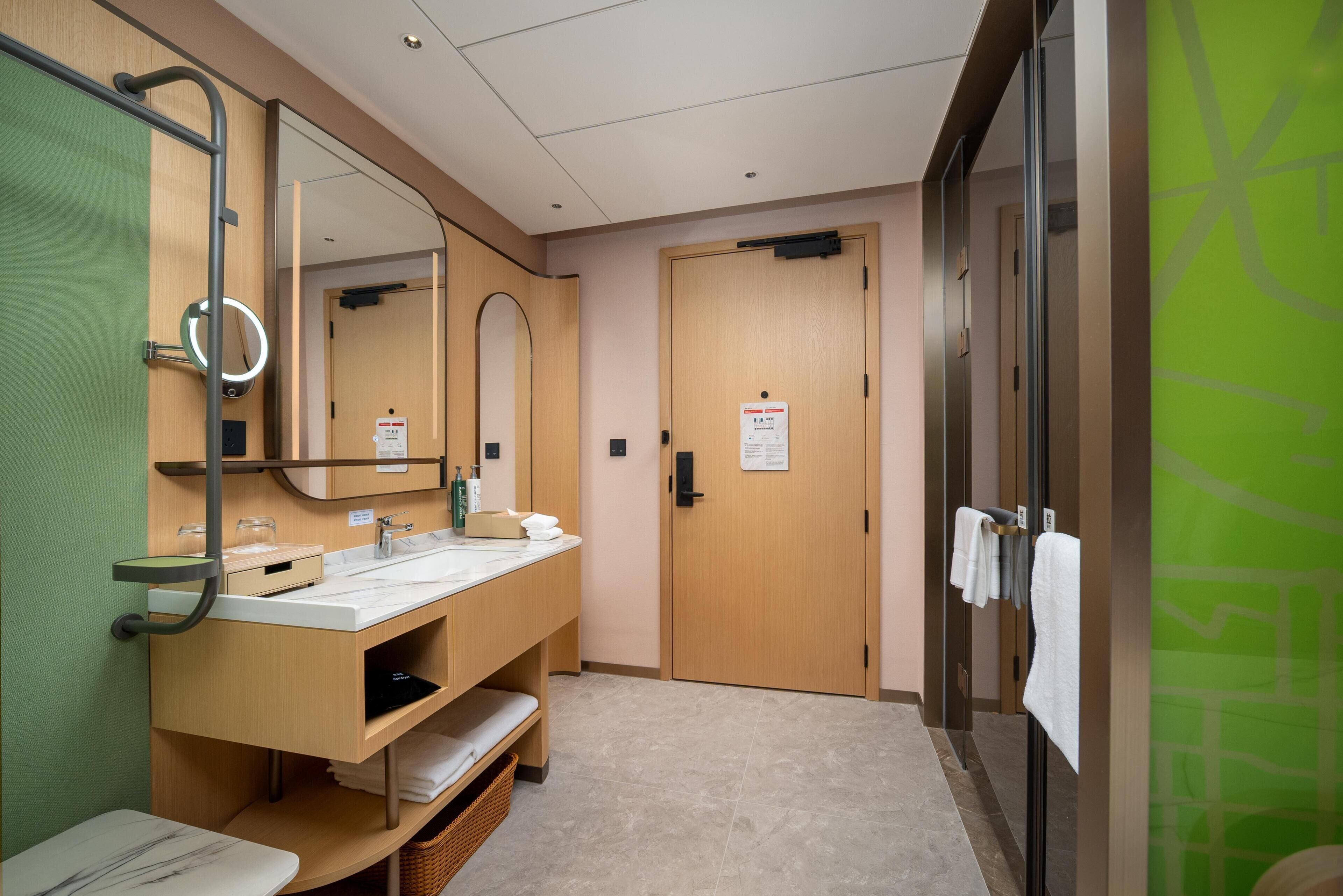 Une salle de bain d’hôtel avec un grand miroir, deux lavabos, un porte-serviettes et une porte en bois.