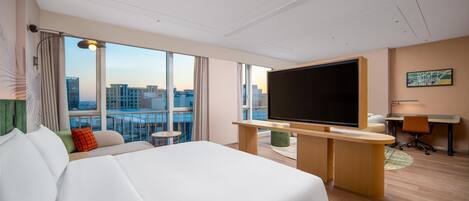A hotel room with a large bed, a desk with a flat-screen TV, a chair, and a view of the city through large windows.