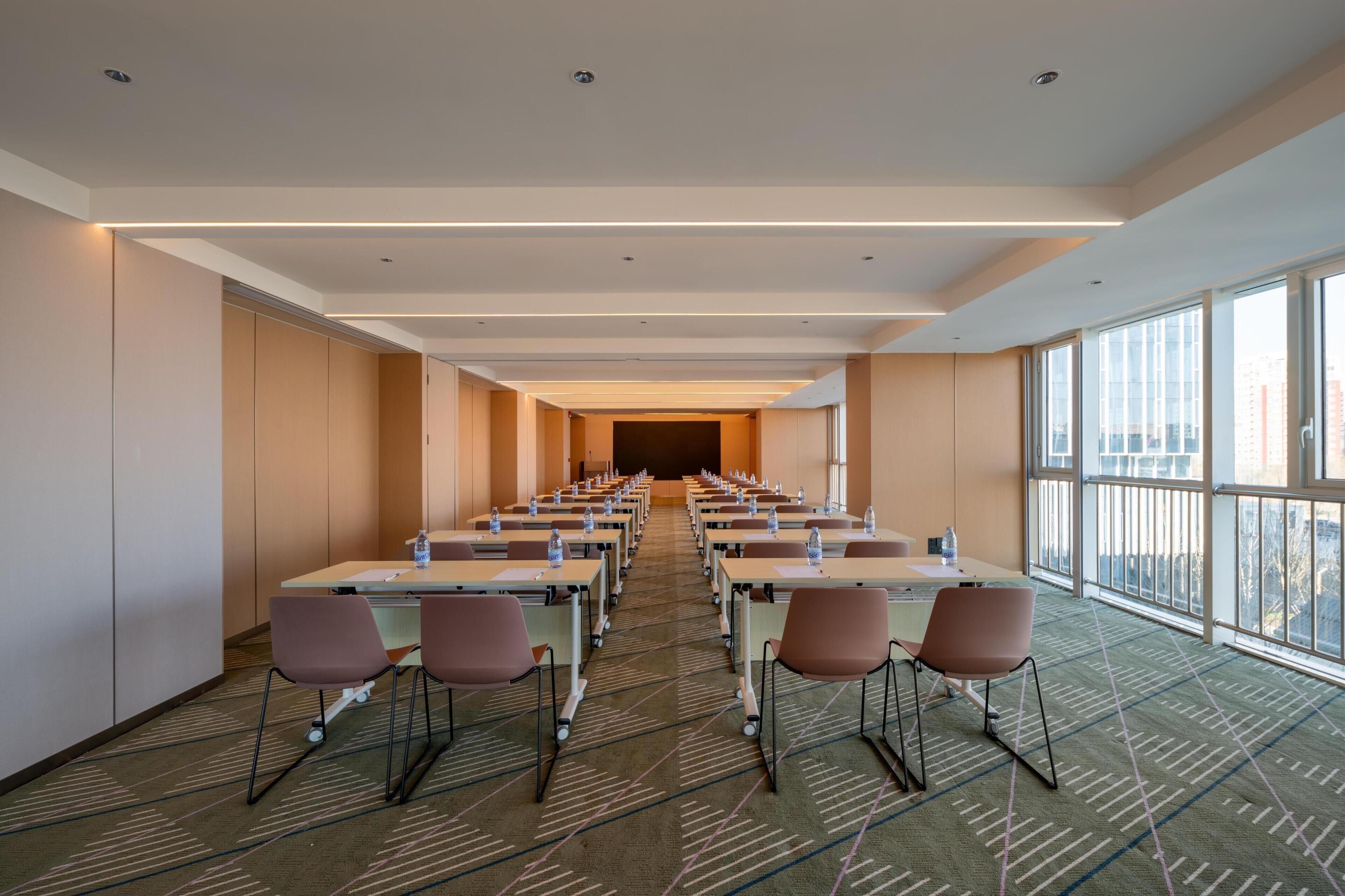 Une salle de conférence avec des rangées de tables et de chaises, de grandes fenêtres et un sol recouvert de moquette.