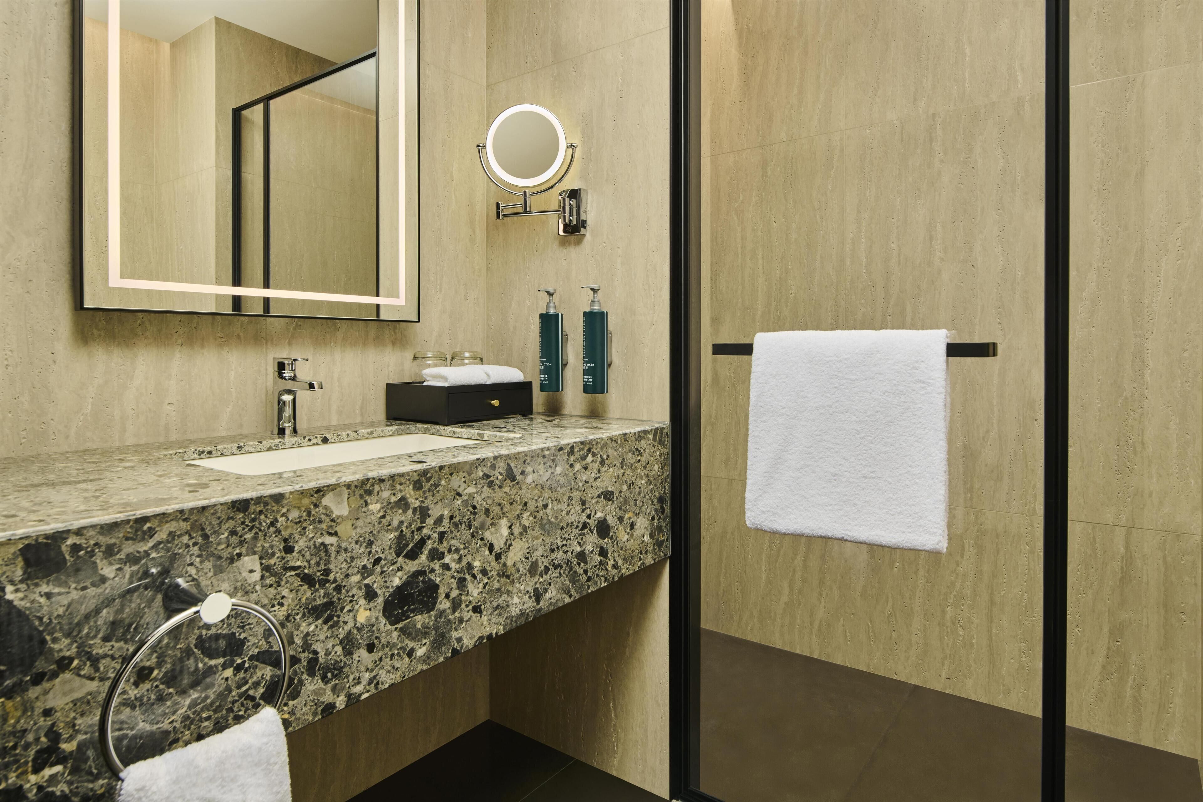 A modern bathroom with a granite countertop, a large mirror, a round mirror, a towel rack, and a glass shower enclosure.