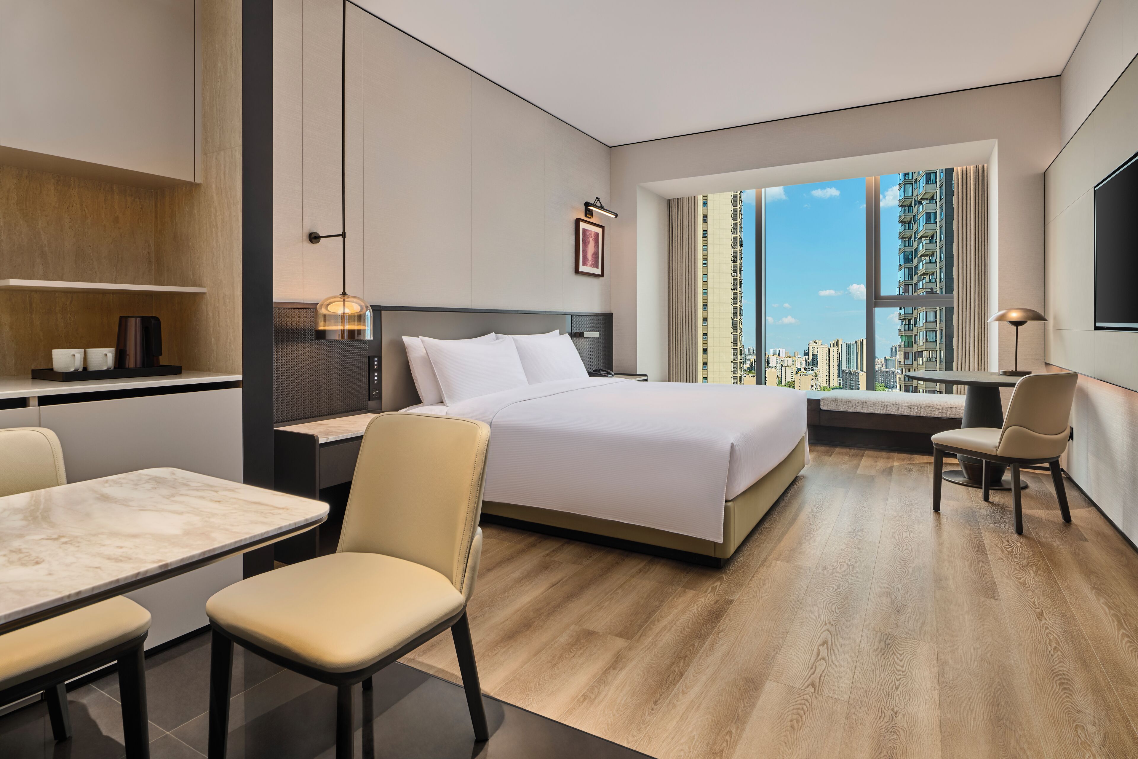 A modern hotel room with a bed, a dining area, and a view of the cityscape.