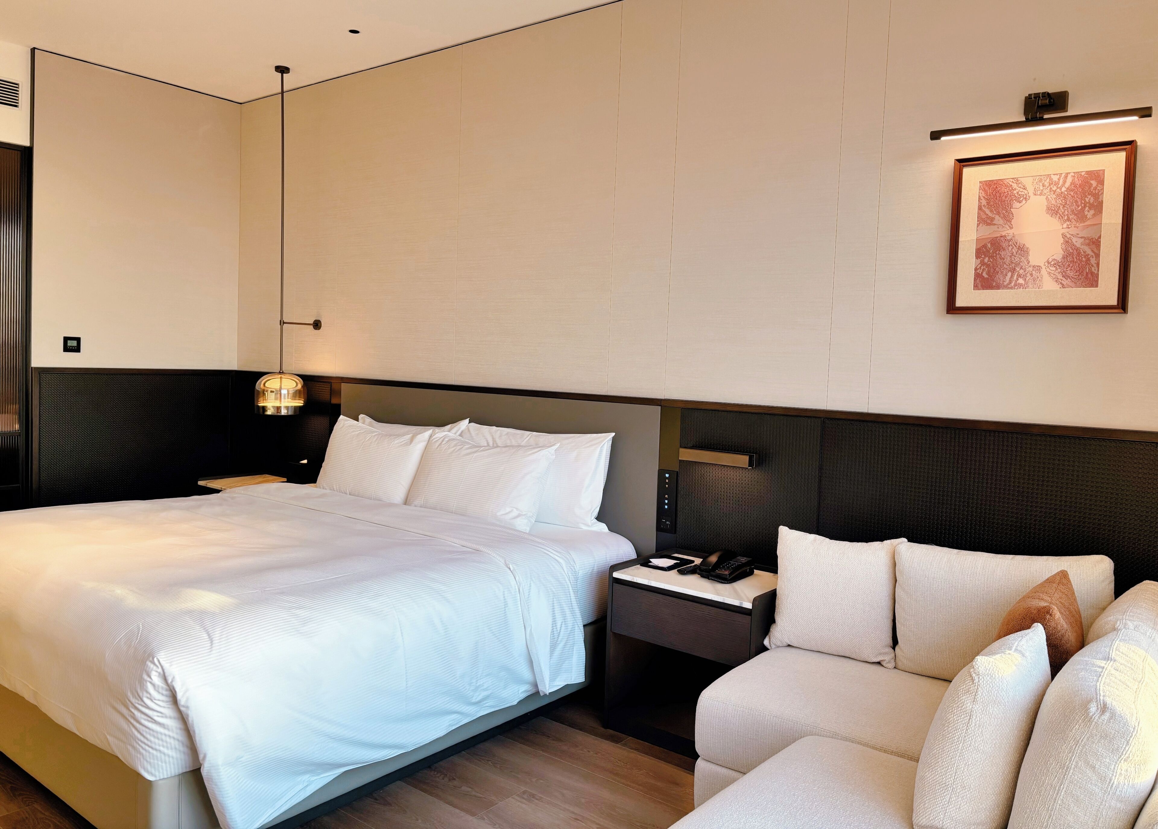 A hotel room with a large bed, a bedside table with a phone and a lamp, a chair with cushions, and a framed picture on the wall.