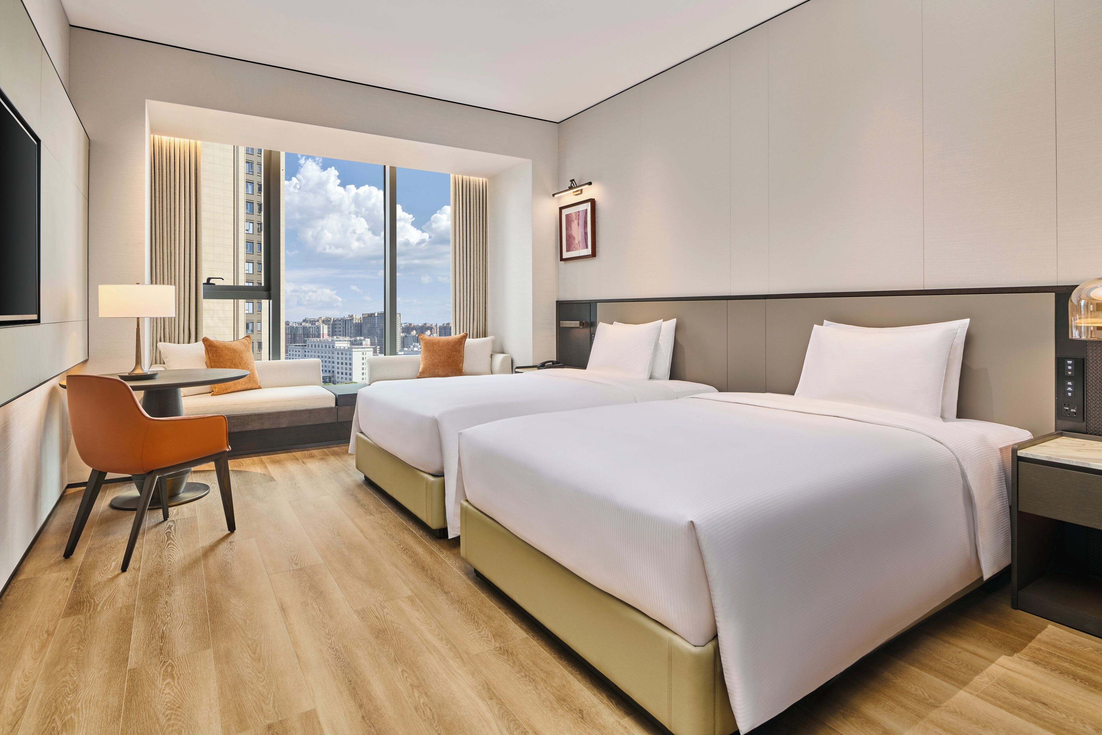 A hotel room with two beds, a sofa, a desk, and a large window with a city view.