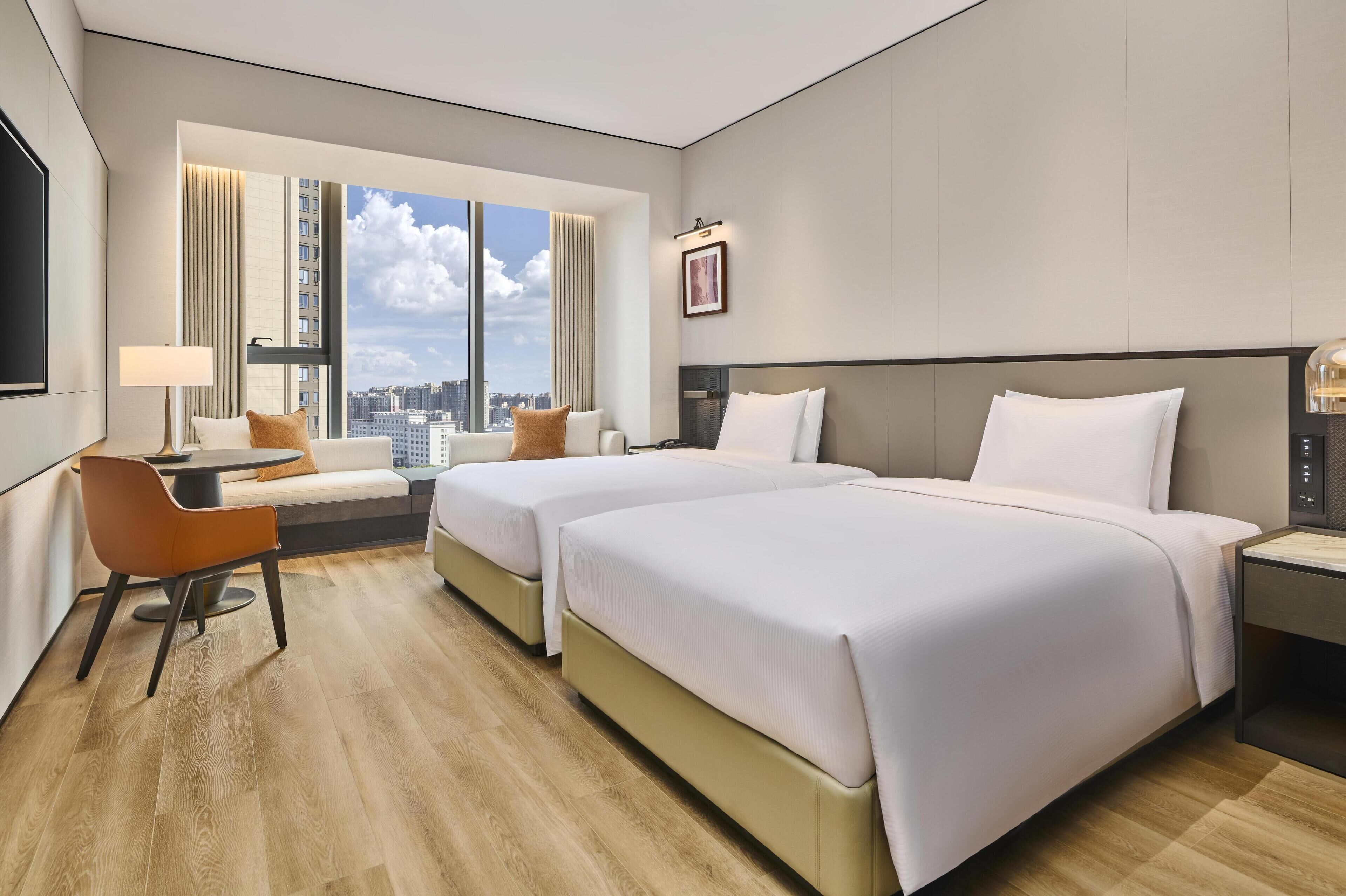 A hotel room with two beds, a desk, a chair, a TV, and a large window with a city view.