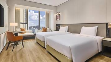 A hotel room with two beds, a desk, a chair, a TV, and a large window with a city view.