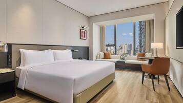 Habitació d'hotel moderna amb un llit gran, un sofà, una cadira i vista a la ciutat a través de grans finestres.