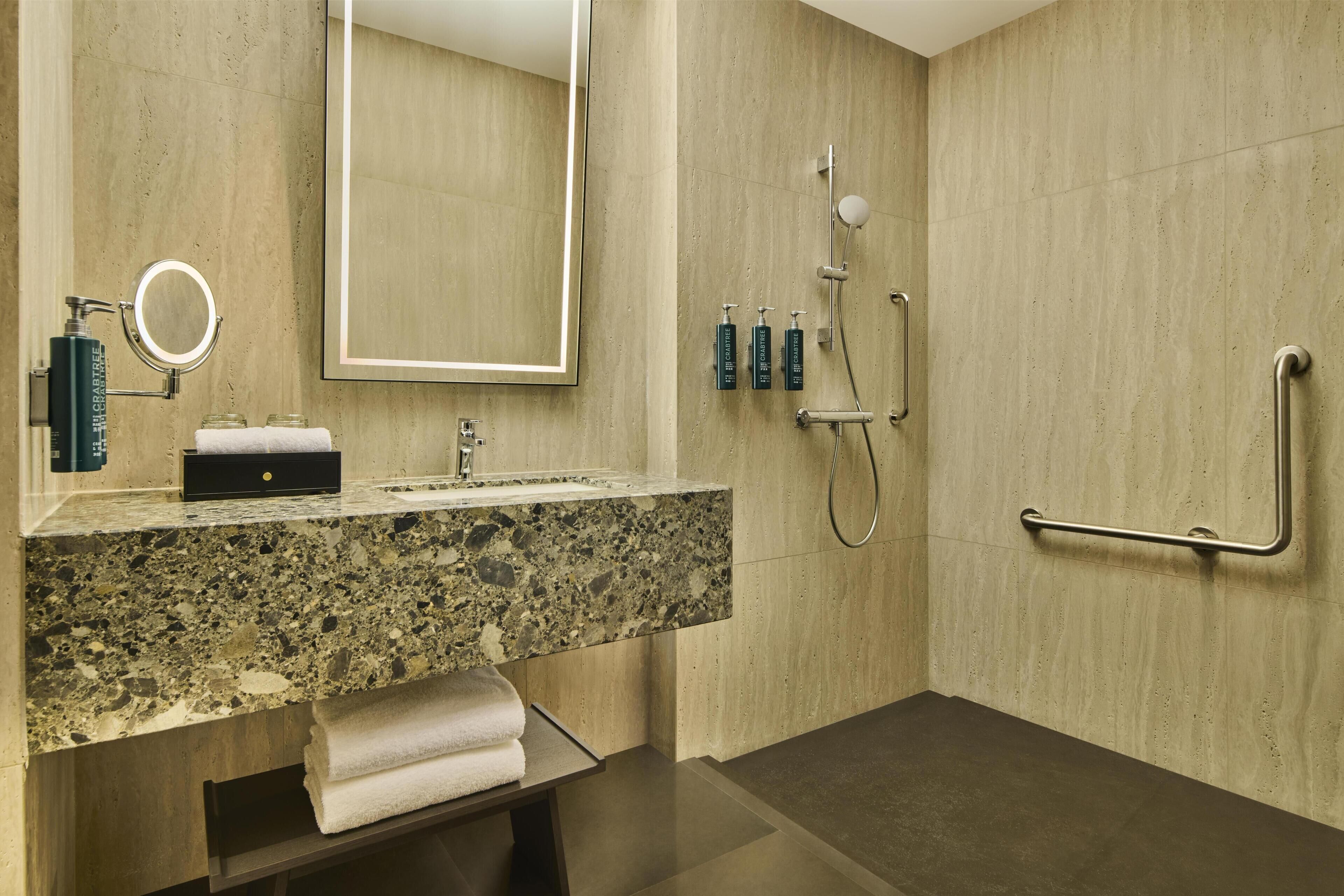 A modern bathroom with a granite countertop, a large mirror, a handheld showerhead, and a grab bar.