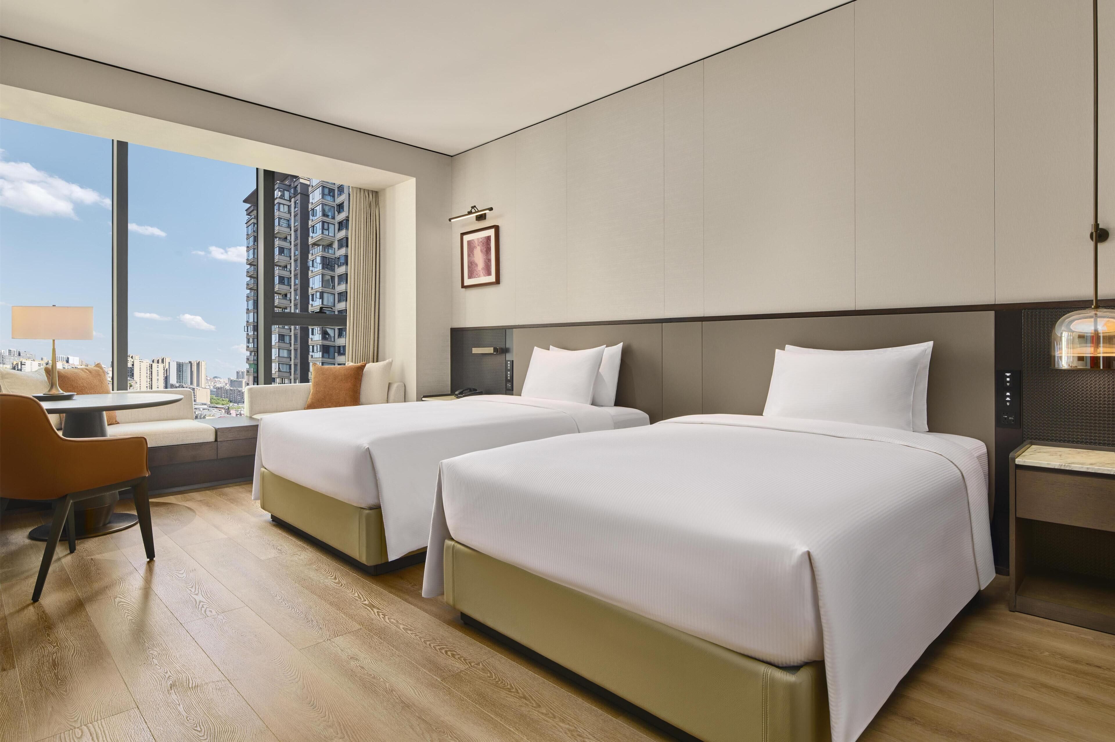 Habitación de hotel con dos camas, un escritorio, una silla y un amplio ventanal con vistas a la ciudad.