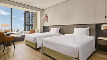 Habitación de hotel con dos camas, un escritorio, una silla y un amplio ventanal con vistas a la ciudad.