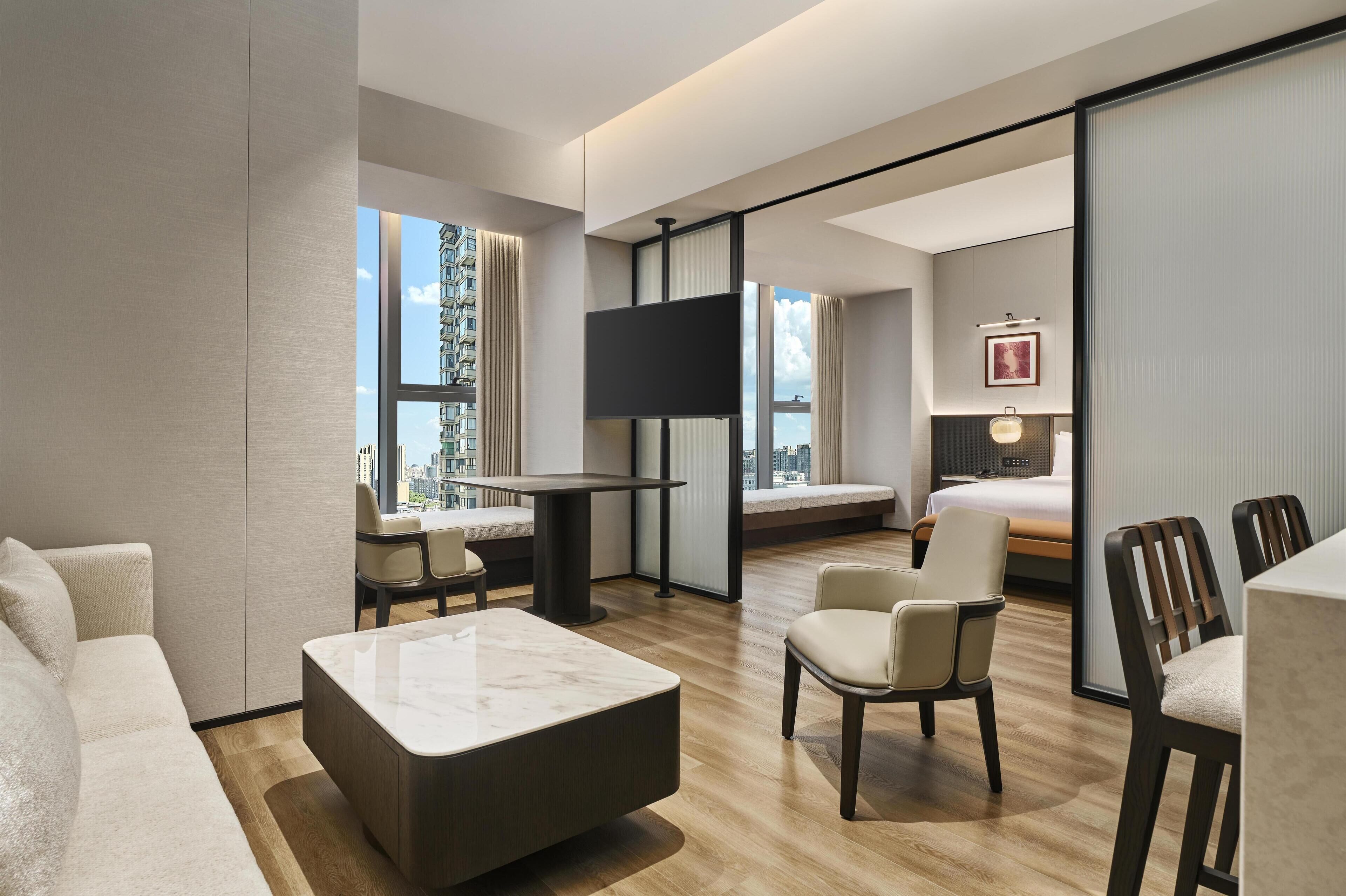 A modern hotel room with a large bed, a TV, a sofa, a coffee table, and a dining area.