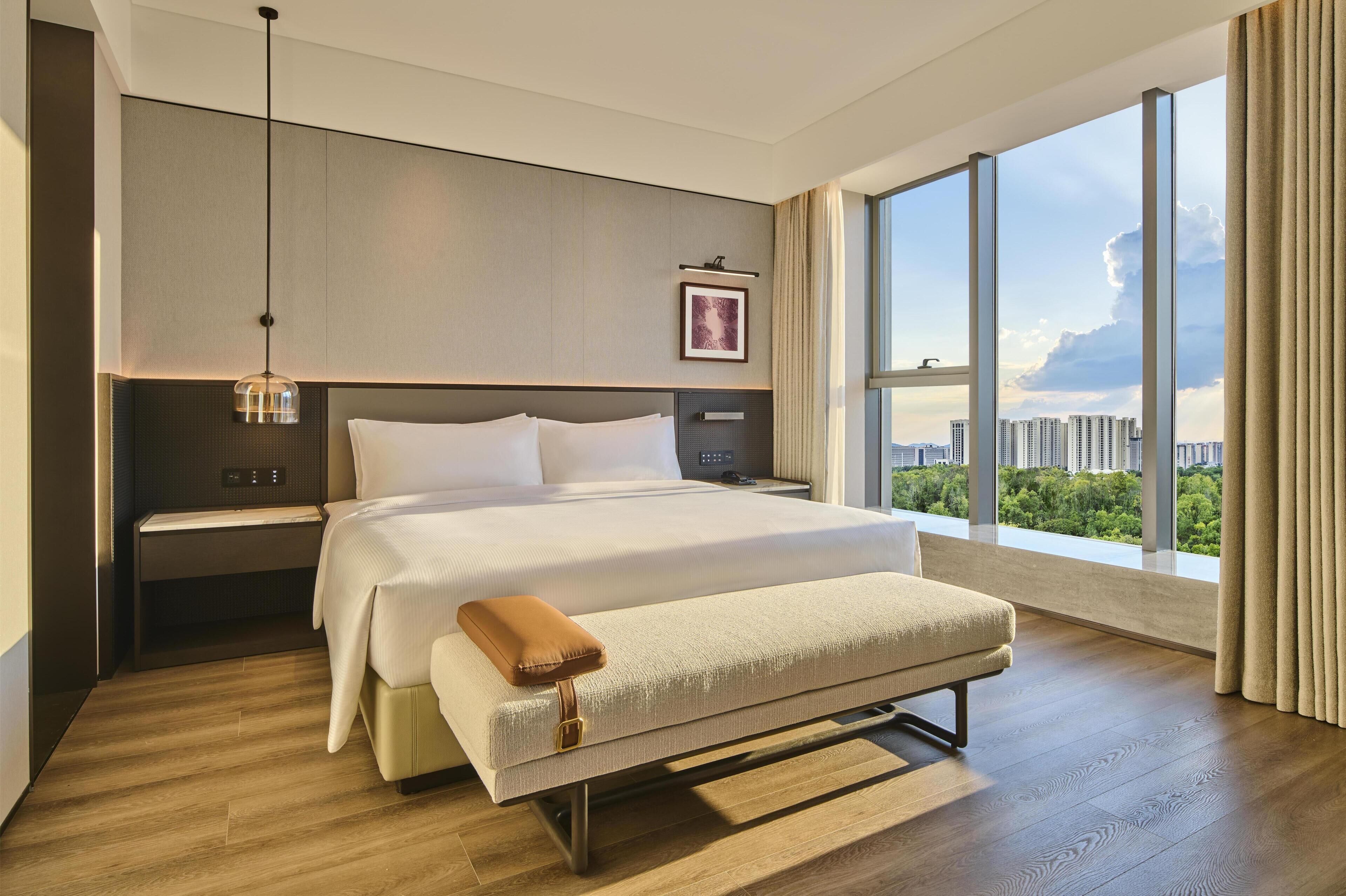 Ein modernes Hotelzimmer mit einem großen Bett, Nachttischen, einer Bank, einer Hängelampe und Blick auf die Stadt durch große Fenster.