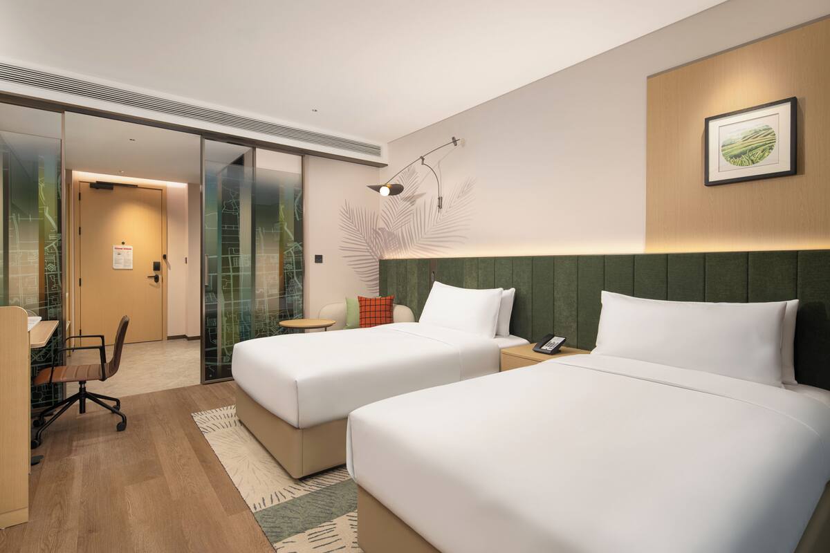 Ein Hotelzimmer mit zwei Betten, einem Schreibtisch, einem Stuhl, einem kleinen Tisch und einer Wandlampe.