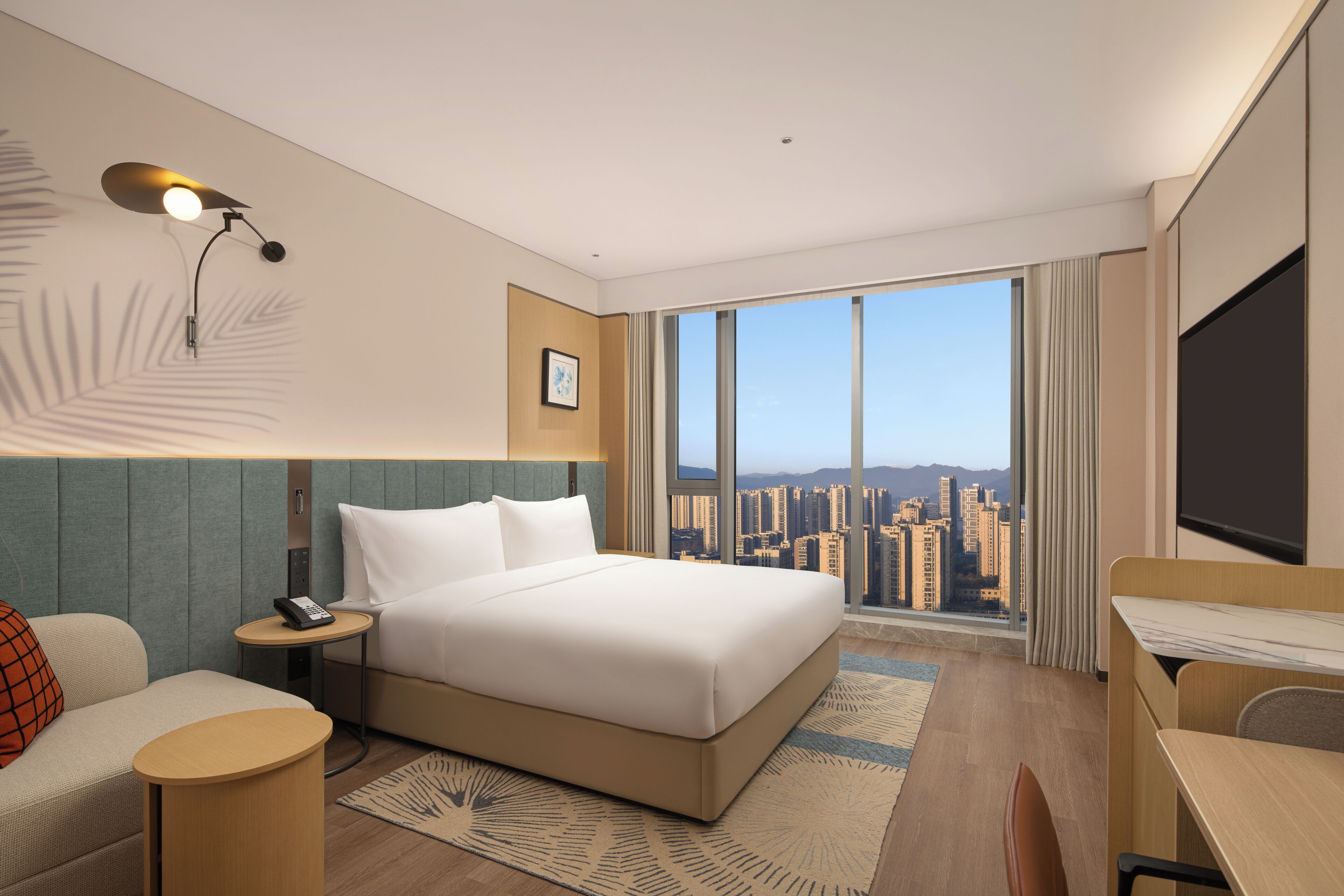 Ein Hotelzimmer mit einem großen Bett, einem Schreibtisch, einem Sessel, einem Nachttisch, einer Lampe, Blick auf die Stadt und einem Fernseher.