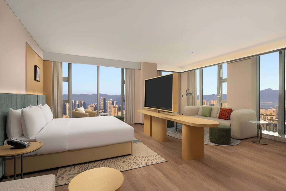 Ein modernes Hotelzimmer mit einem großen Bett, einem Fernseher, einer Sitzecke und Blick auf die Stadt.