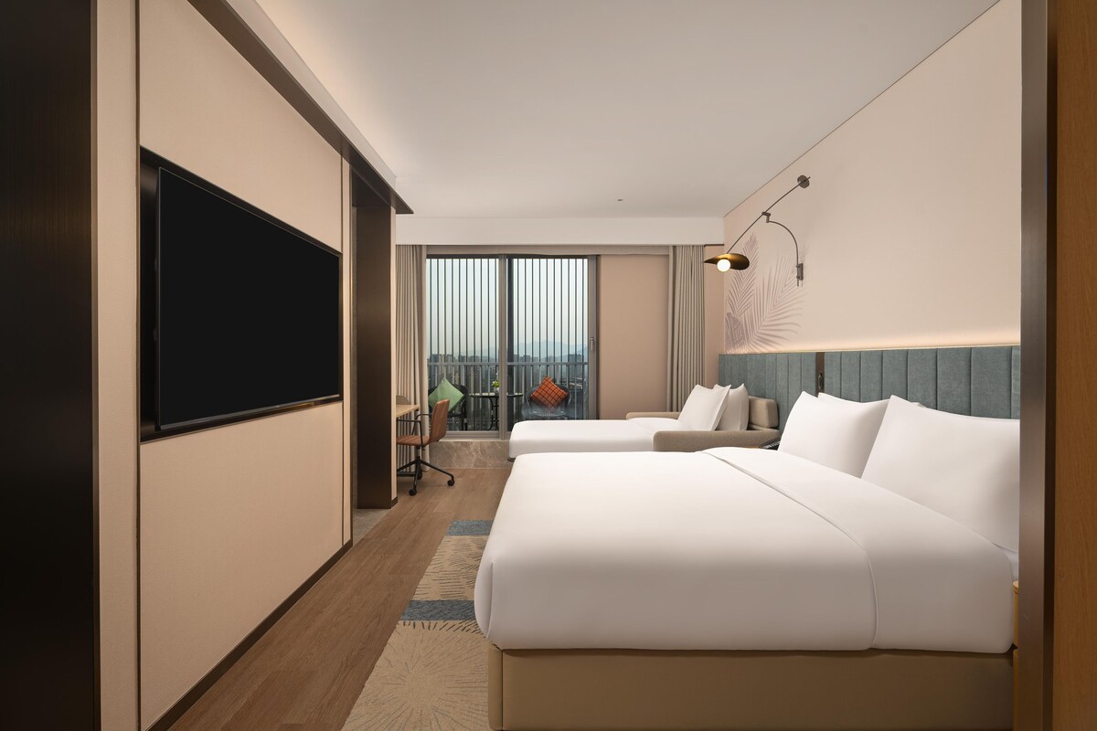 A hotel room with two beds, a TV, a desk, and a balcony with a view.