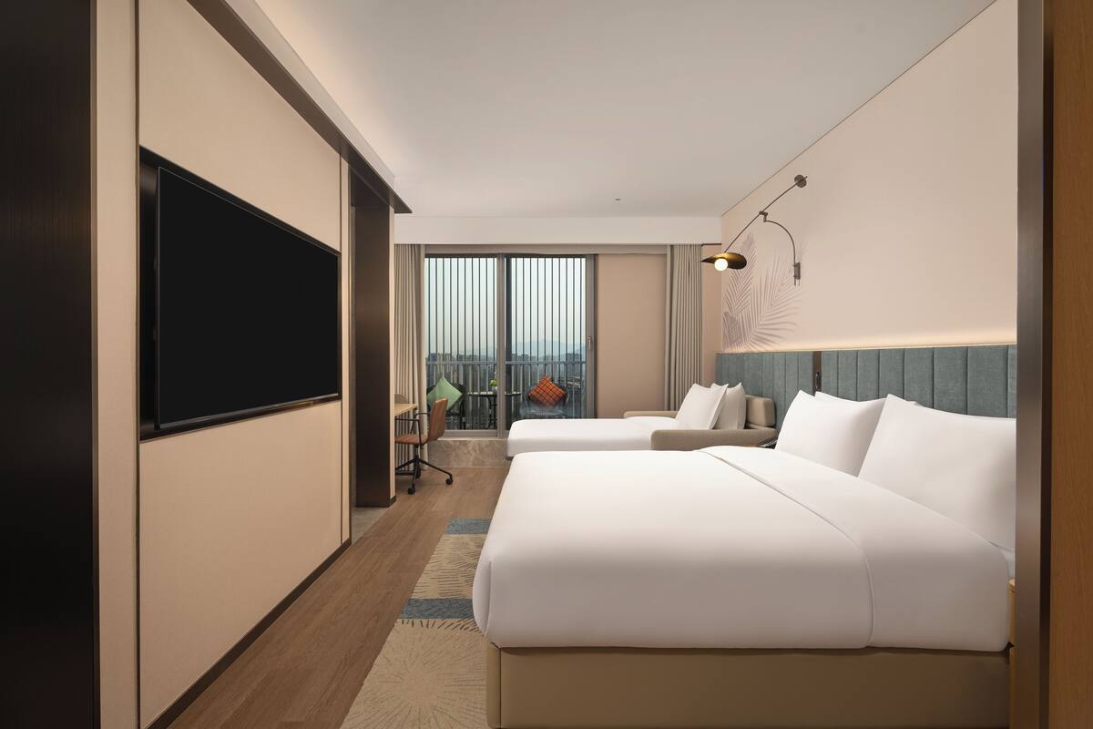 Ein Hotelzimmer mit zwei Betten, einem Fernseher, einem Schreibtisch und Blick auf die Stadt.