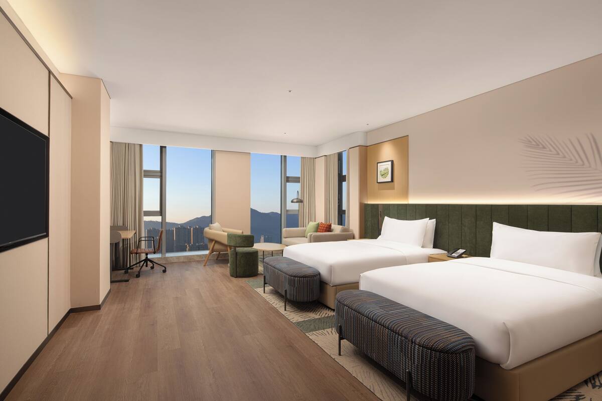 Ein Hotelzimmer mit zwei Betten, einem Schreibtisch, einem Sessel und einem Sofa, sowie einem großen Fenster mit Blick auf die Stadt.