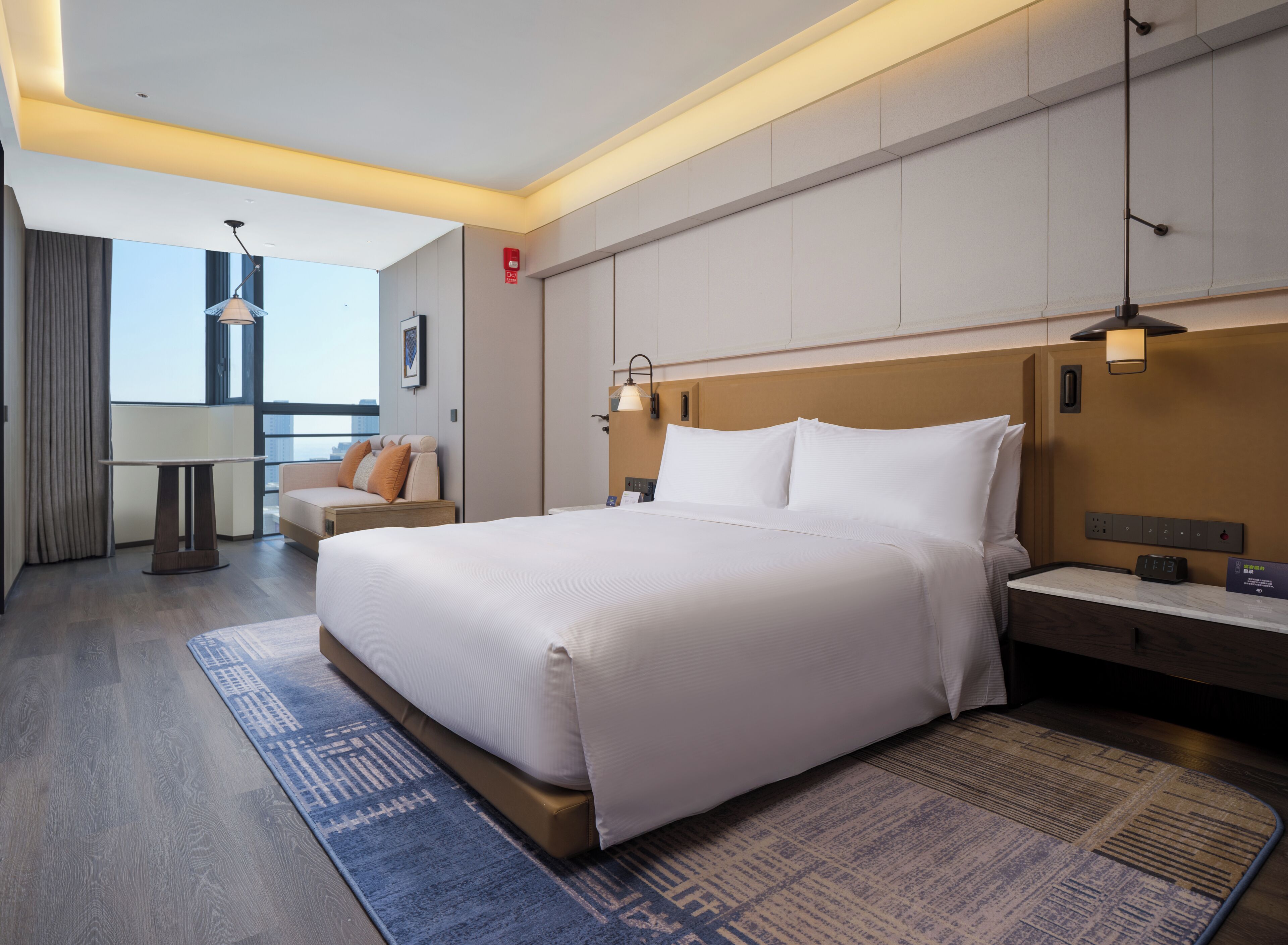 Quarto de hotel com cama grande, mesa de cabeceira, abajur, cortinas e vista da cidade.