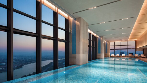 Piscina interior con amplios ventanales que ofrecen vistas a la ciudad, diseño moderno con techos altos y sillones de descanso junto a la piscina.