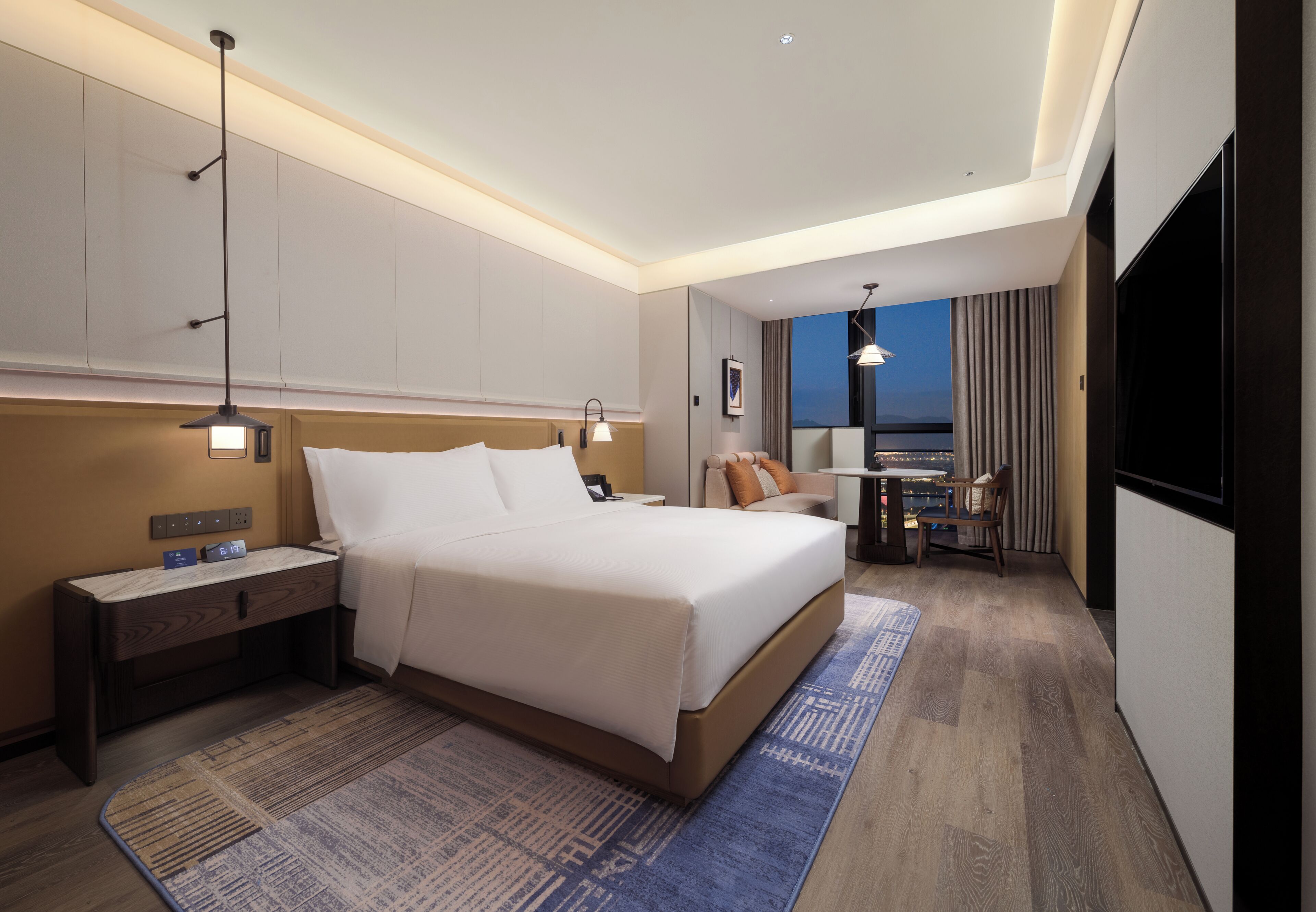 Quarto de hotel com cama, mesa de cabeceira, abajur e vista para a cidade.