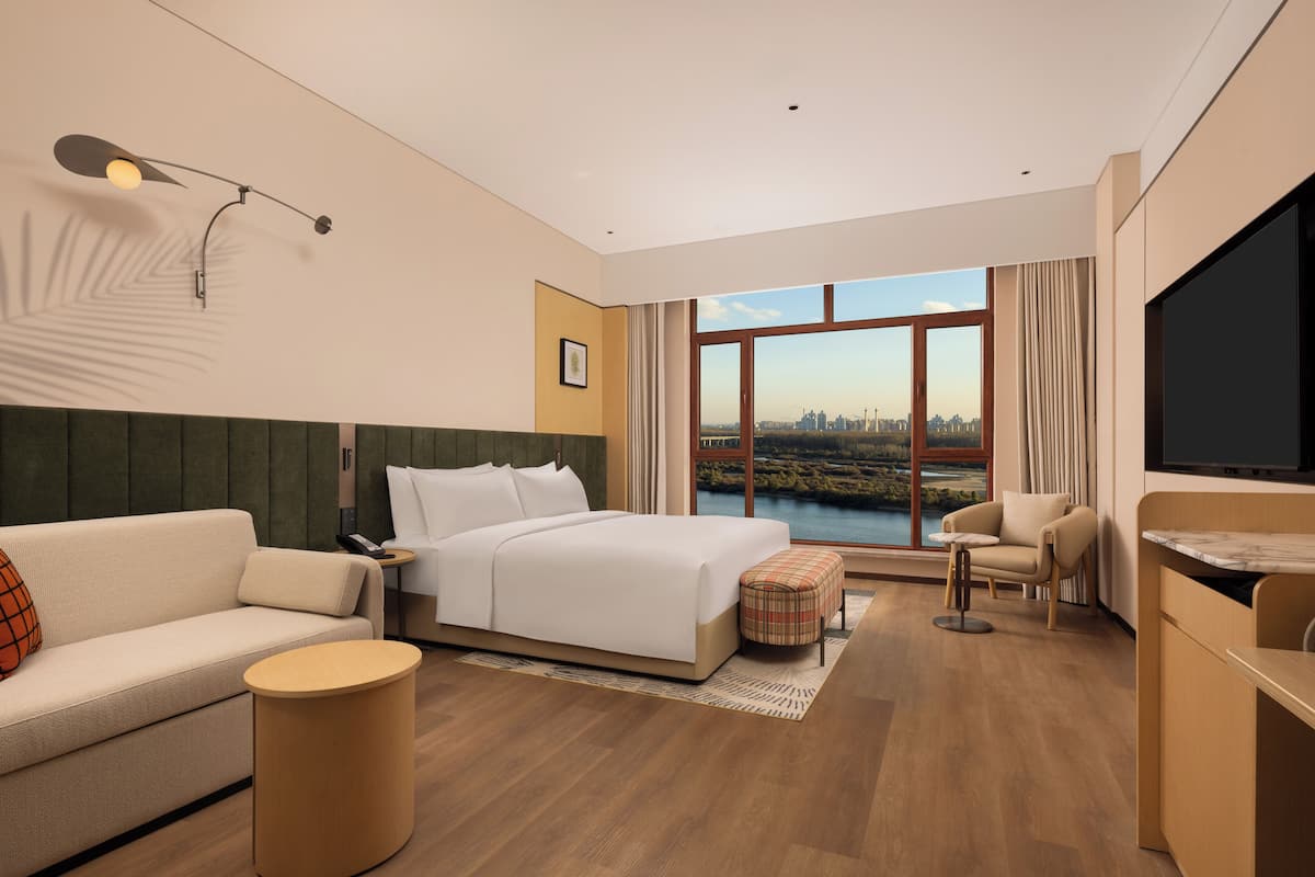 A modern hotel room with a large bed, a sofa, a small round table, a chair, and a TV.