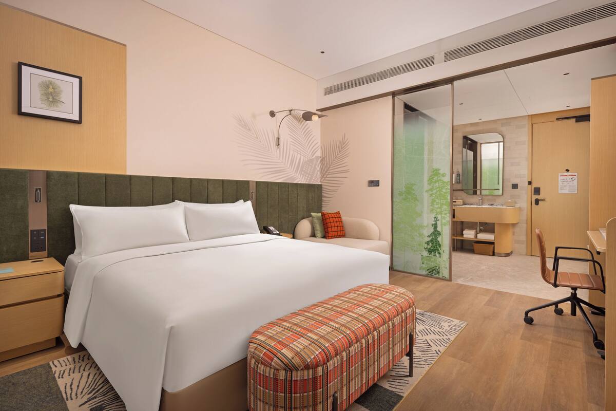 A modern hotel room with a large bed, a bench, a desk, and a bathroom visible through an open door.