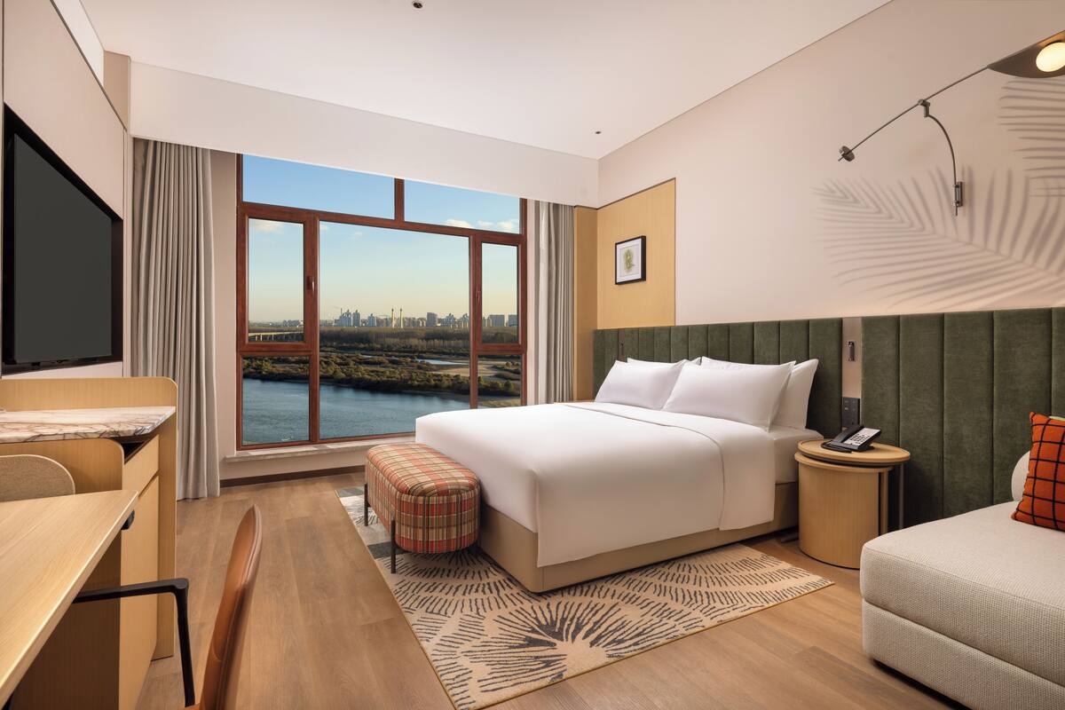 A hotel room with a large bed, a desk, a chair, a small table, a bedside table, and a view of a cityscape through large windows.