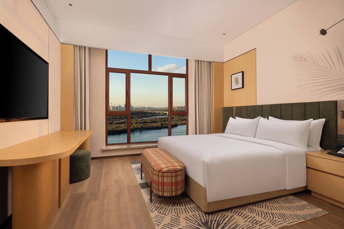 A modern hotel room with a large bed, a desk, a television, and a view of a cityscape through large windows.