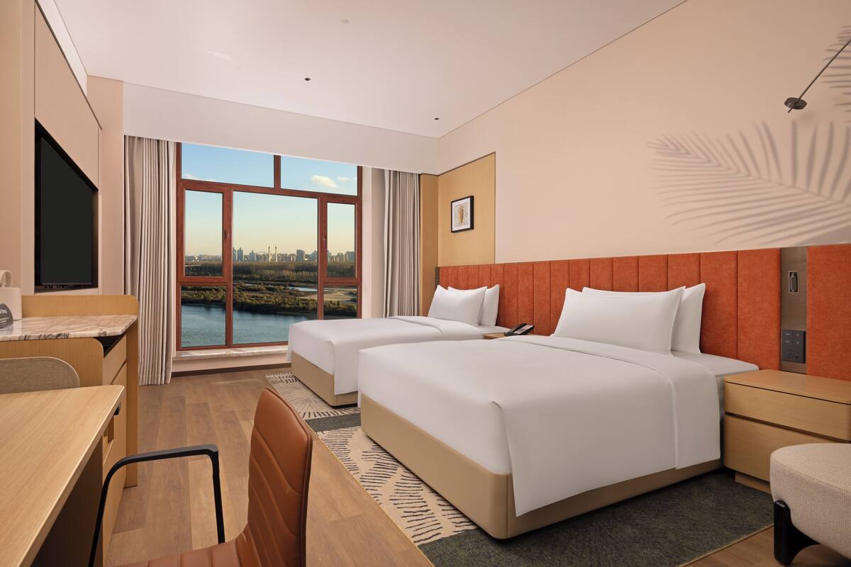 A hotel room with two beds, a desk, a chair, and a large window with a city view.