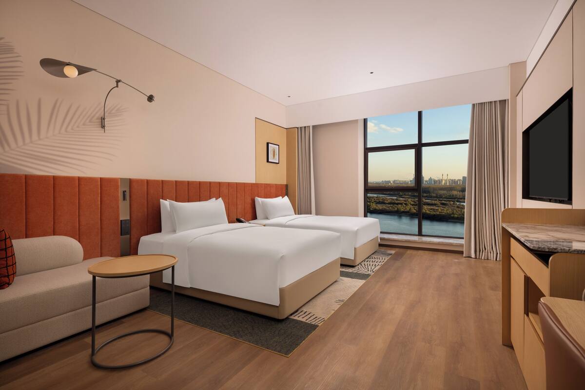 A modern hotel room with two beds, a sofa, a small table, and a large window offering a city view.