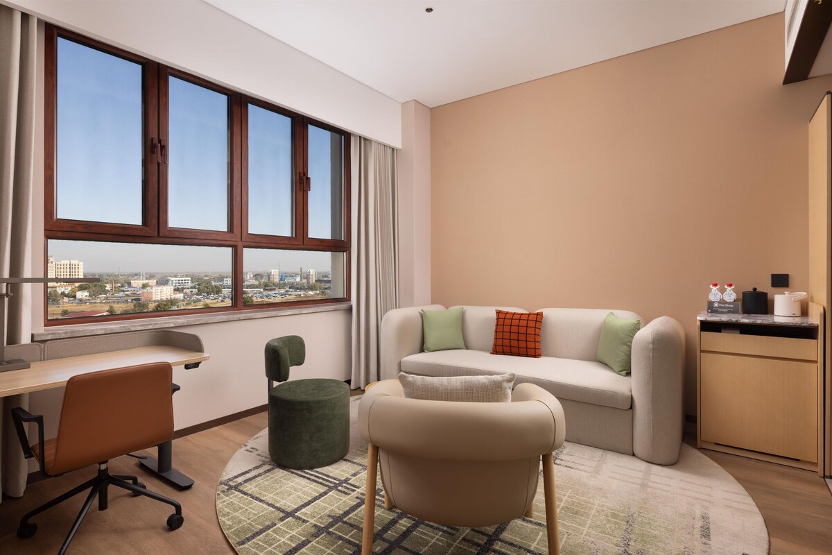 A modern living room with a large window, a sofa, a desk, and a green ottoman.