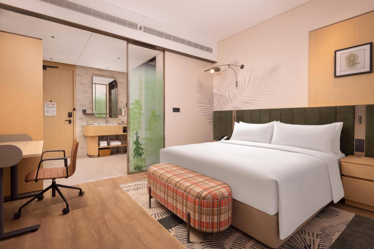 A modern hotel room with a large bed, a desk with a chair, a bench, a large mirror, and a wall-mounted light fixture.