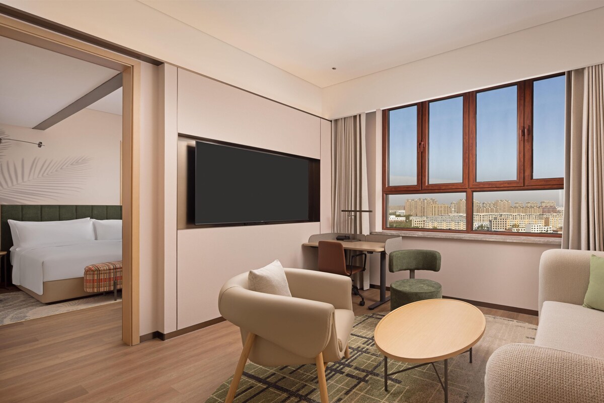 Ein modernes Hotelzimmer mit Bett, Fernseher, Essbereich und großem Fenster mit Blick auf die Stadt.