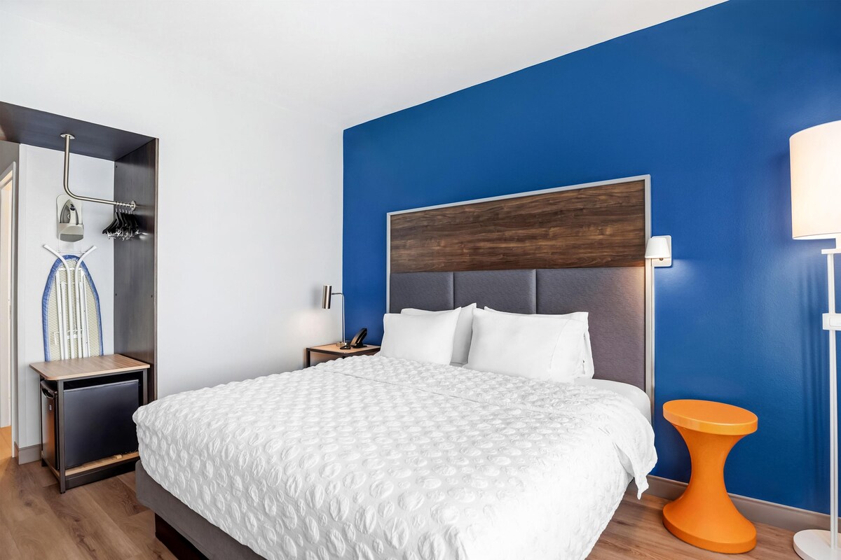 Ein modernes Hotelzimmer mit einem großen Bett, einem Nachttisch, einer Wandlampe und einem auffälligen orangefarbenen Hocker.
