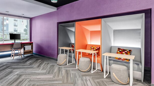 Une pièce moderne avec un mur violet, un bureau avec un ordinateur, deux tables et un canapé avec des coussins.