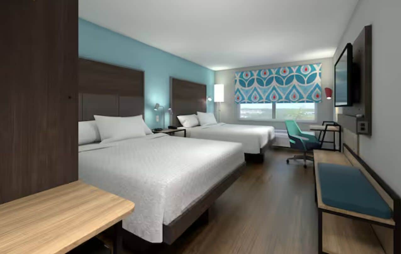 A hotel room with two beds, a desk, a chair, a TV, and a window with a view.