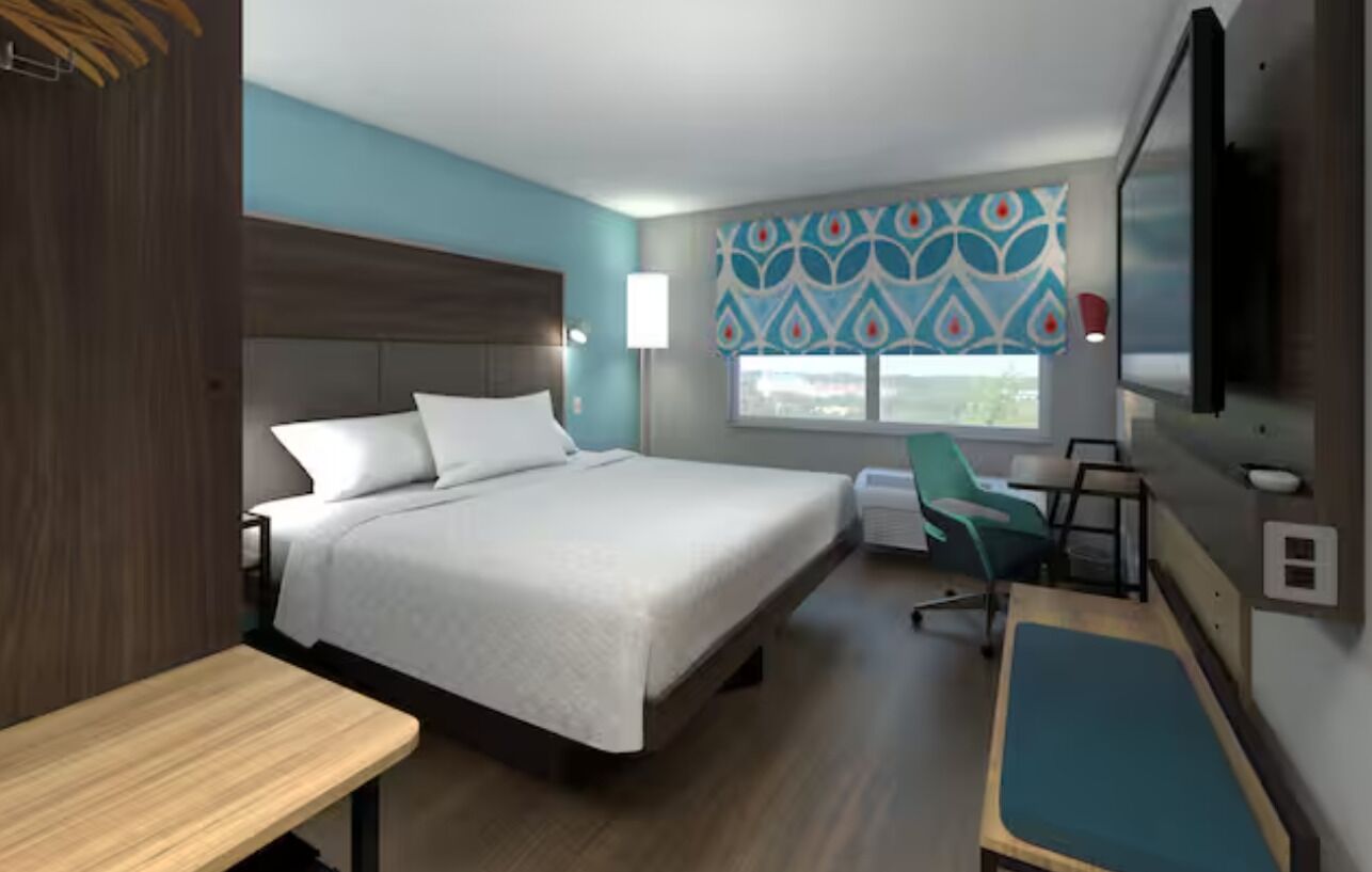 A hotel room with a bed, a desk, a chair, a TV, and a window with patterned curtains.