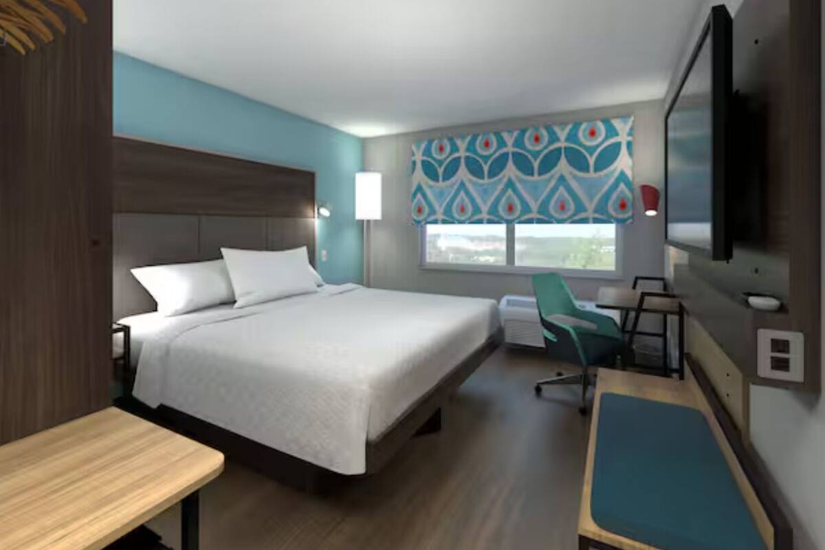 Ein Hotelzimmer mit Bett, Schreibtisch, Stuhl, Fernseher und einem Fenster mit gemusterten Vorhängen.