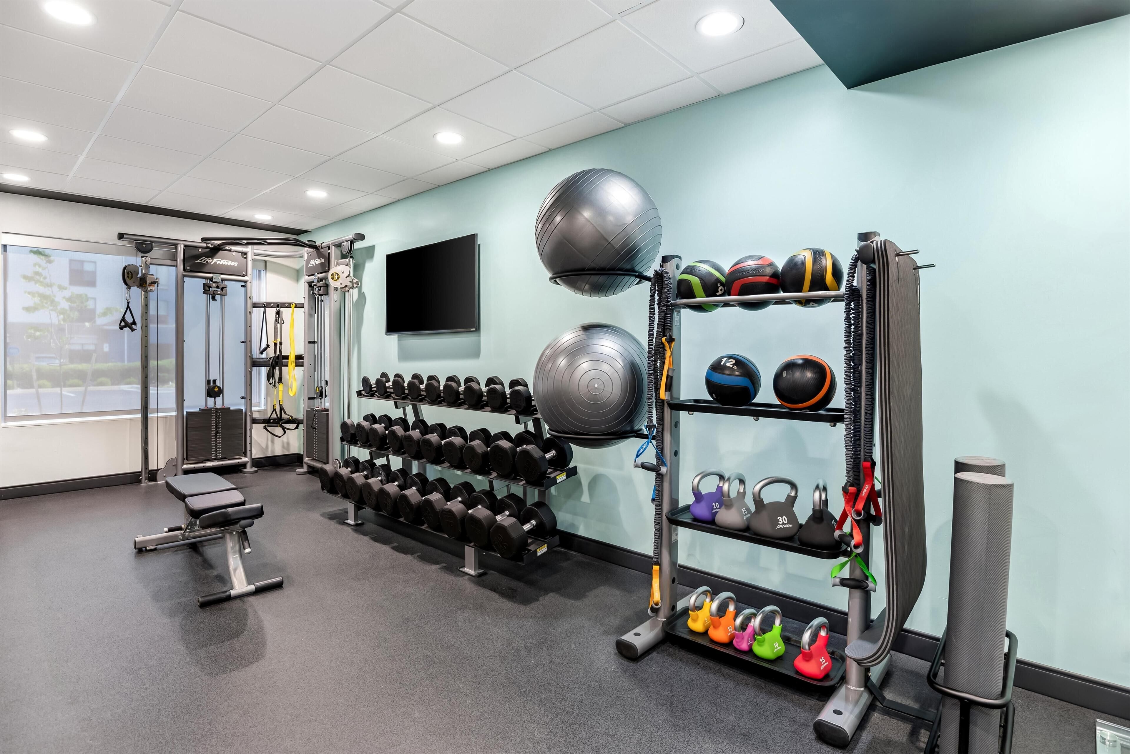 Un gimnasio con equipo de levantamiento de pesas, kettelbells y un televisor en la pared.